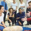 a mixed age group listen to their tutor as he shows the procedure involved to resuscitate using a defibrillator .